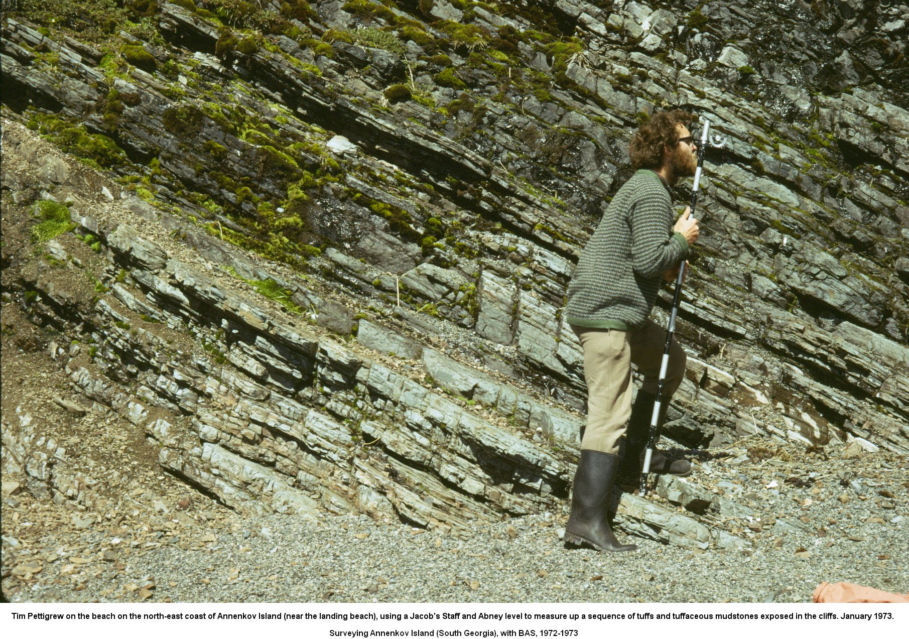 Tim Pettigrew on the beach on the north-east coast of Annenkov Island (near the landing beach), using a Jacob's Staff and Abney level to measure up a sequence of tuffs and tuffaceous mudstones exposed in the cliffs. January 1973.