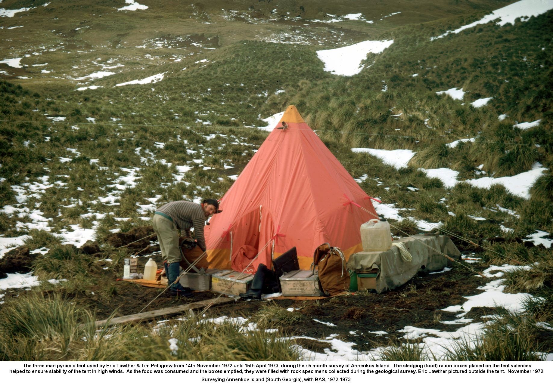 The three man pyramid tent used by Eric Lawther & Tim Pettigrew from 14th November 1972 until 15th April 1973, during their 5 month survey of Annenkov Island.  The sledging (food) ration boxes placed on the tent valences
helped to ensure stability of the tent in high winds.  As the food was consumed and the boxes emptied, they were filled with rock specimens collected during the geological survey. Eric Lawther pictured outside the tent.  November 1972.